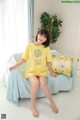 A woman sitting on a bed wearing a yellow shirt and shorts.