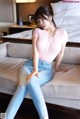 A woman sitting on a couch wearing a pink shirt and jeans.